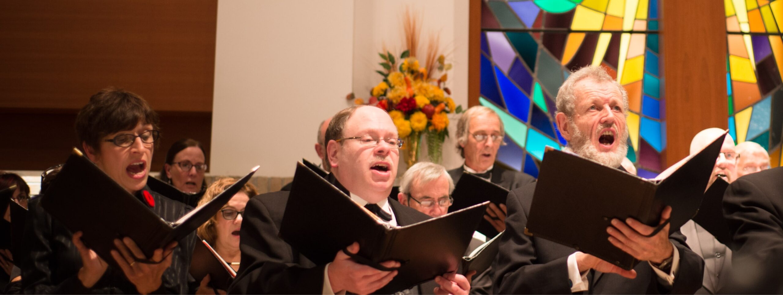 Closeup of three people singing in a choir in front of stained glass windows